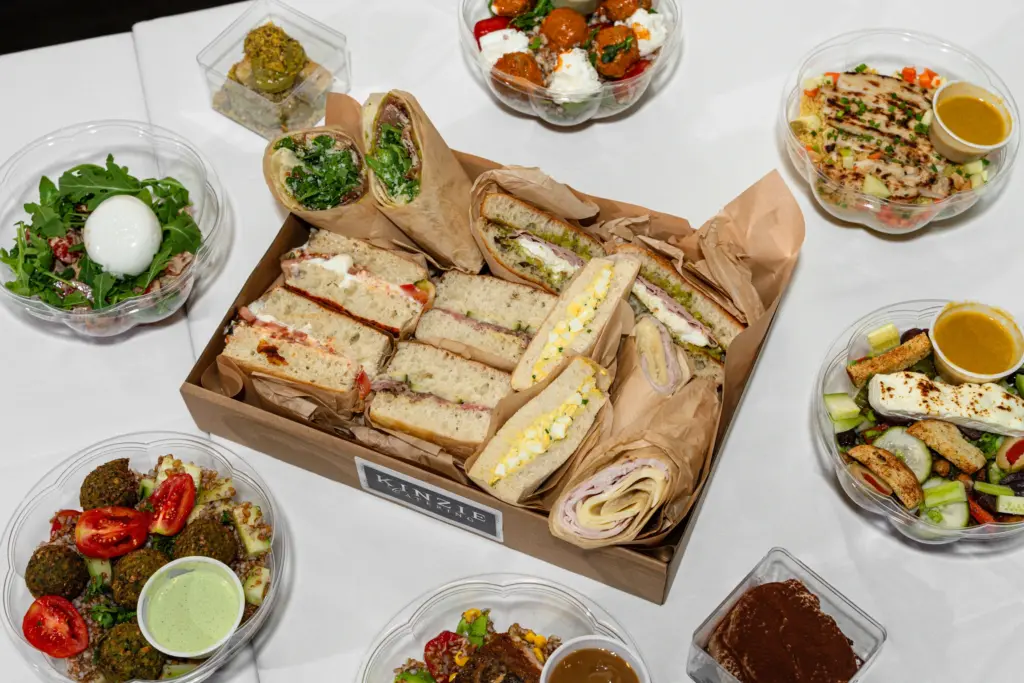A top-down view of a catering spread on a white tablecloth, featuring a central wooden tray with assorted sandwiches and wraps. Surrounding the tray are several clear plastic bowls containing fresh salads, falafel with tahini sauce, and grilled chicken skewers. The arrangement suggests a well-prepared and appetizing meal for an event or gathering.