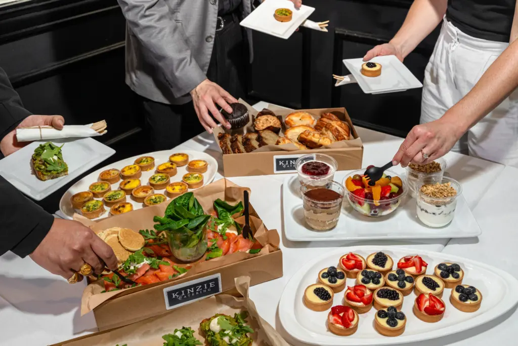 A top-down view of a catered event setup, featuring multiple food stations on a white tablecloth. Several people are serving themselves from the spread: one person is taking a slice of layered vegetable tart, another is selecting a chocolate cupcake from a box labeled “KINZIE,” and a third is scooping salad into a bowl. The table displays an array of gourmet appetizers, including mini quiches with black olives and strawberries, individual dessert cups with granola, and a colorful fruit salad. In the background, two waiters hold plates of canapés, ready to serve guests.
