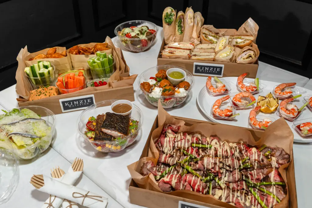 An overhead view of a diverse catering spread on a white tablecloth, featuring multiple food stations. In the foreground, a large cardboard box holds strips of steak with asparagus and a creamy sauce drizzle. To the left, there’s a glass bowl of salad with fish fillets and another of crab cakes. A central platter displays shrimp cocktail and a bowl of stuffed grape leaves with dipping sauce. In the background, a box contains sandwiches and wraps, alongside cups of celery sticks, carrot sticks, and cucumber rounds. The scene is branded with “KINZIE CATERING” labels on the boxes, set against a dark background for emphasis.